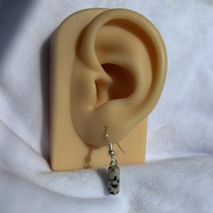 Pode incluir: Um &uacute;nico brinco pendurado com uma conta cil&iacute;ndrica de pedra salpicada. A conta &eacute; bege com manchas pretas, suspensa por um gancho prateado. O brinco &eacute; exibido em uma orelha de manequim contra um fundo branco.