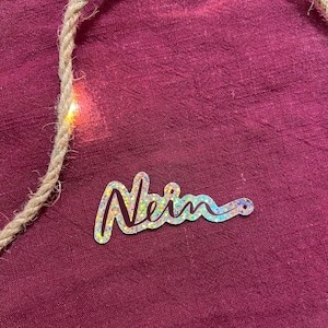 Könnte beinhalten: Das Wort "Nein" in glitzerndem Silber auf einem burgunderfarbenen Stoffhintergrund.