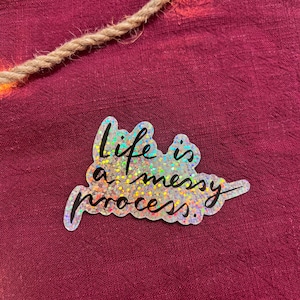 Könnte beinhalten: Ein silberner holografischer Aufkleber mit dem Text "Life is a messy process." in schwarzer Schrift.