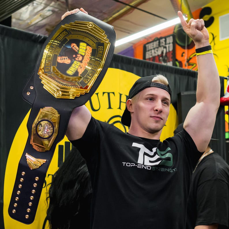 May include: A man wearing a black t-shirt with the text "TOP-END STRENGTH" holds a gold and black championship belt with the text "FEMALE ESTER BEAT DOWN" and "SOME 9999 WR".