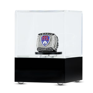 May include: A silver championship ring with a blue and red shield featuring the letters "FFL" and the words "League Champion". The ring is displayed in a clear acrylic case with a black base.