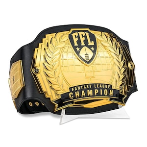 May include: A black and gold championship belt with the text "FFL FANTASY LEAGUE CHAMPION". The belt features a gold center plate with a football and laurel wreath design. The belt is displayed on a clear stand.