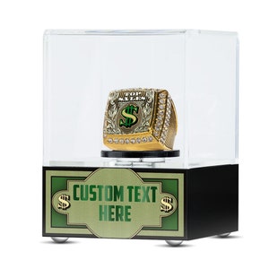 May include: A gold-colored ring with green and white accents, featuring a dollar sign and the words "100 Trillion Dollars" on the top. The ring is displayed in a clear acrylic case with a black base. The base has a green and gold label with the text "CUSTOM TEXT HERE" and dollar signs.