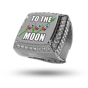 May include: A silver ring with a large square face featuring the text "TO THE MOON" with a green arrow pointing up and three cartoon rockets. The ring is encrusted with clear rhinestones.