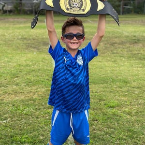 May include: A young boy wearing a blue and black striped soccer jersey and blue shorts holds a black championship belt above his head. The belt has gold accents and says "Player of the Game" and "FCG Futbol Club Gators".