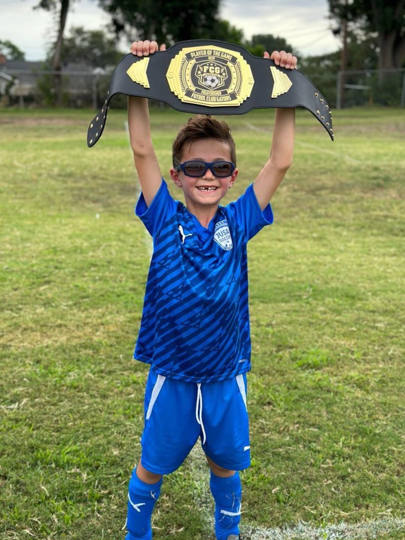 May include: A young boy wearing a blue and black striped soccer jersey and blue shorts holds a black championship belt above his head. The belt has gold accents and says "Player of the Game" and "FCG Futbol Club Gators".