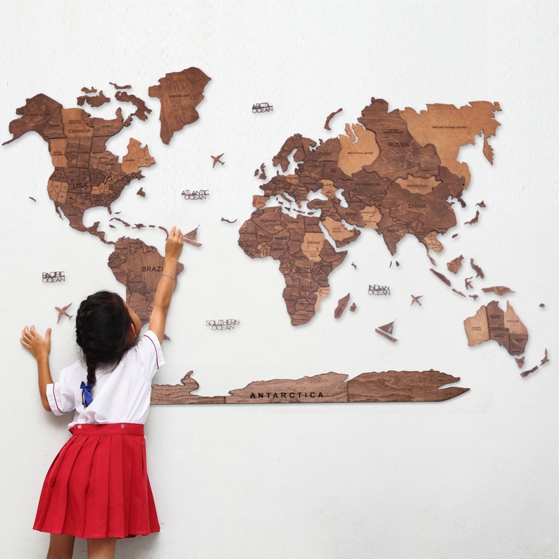 May include: A detailed wooden world map, featuring country names, mounted on a white wall. The map is crafted from various shades of brown wood. Small wooden airplane cutouts are placed around the map. A person is reaching towards the map.