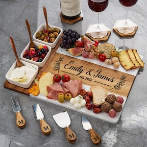 May include: A rectangular wooden and white marble serving board engraved with the text Emily & James est. 2022, featuring three square white ceramic bowls with wooden spoons, four metal cheese knives with wooden handles, and two wine glasses.