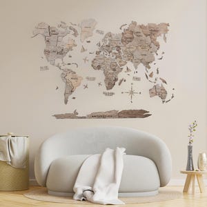 May include: A wooden world map with a compass and airplane cutouts. The map is mounted on a light beige wall above a white sofa with a white throw blanket.