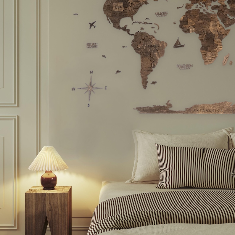 May include: A wooden world map with a compass and the words "USA", "Brazil", "Atlantic Ocean", "Southern Ocean", and "Antarctica" on it. The map is hanging on a wall above a bed with a white headboard and a striped duvet.