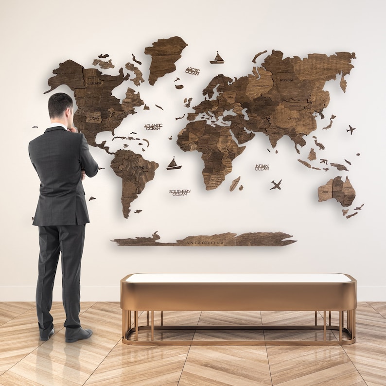 May include: A large wooden world map fixed to a white wall. The map is made of dark brown wood and shows detailed country outlines. A person in a suit stands in front of the map, contemplating it. A gold bench with a white top is in the foreground.