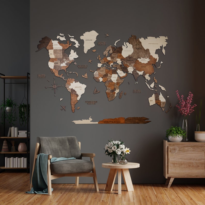 May include: A wooden world map wall decor in light brown and beige tones. The map is mounted on a grey wall and features a compass, aeroplanes, and sailboats. The map is surrounded by a grey wall, a wooden side table, a grey armchair, and a vase of white flowers.
