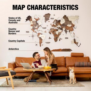 May include: A wooden world map with a light brown background and dark brown borders. The map shows the states of the US, Canada, Australia, and country names and borders. The map is hanging on a white wall in a living room with a brown leather sofa and a dog.