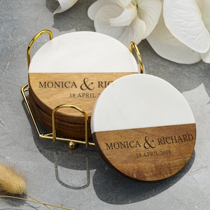 May include: A set of personalized coasters with a gold-colored metal holder. Each coaster is round, with a white top half and a wooden bottom half. The text "MONICA & RICHARD" and the date "18 APRIL 2023" are printed on the wooden section.