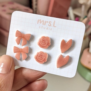 Può includere: Un set di sei orecchini a bottone color pesca a forma di fiocchi, rose e cuori. Gli orecchini sono esposti su una carta bianca con la scritta "mrs.L studio".