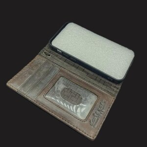 May include: A brown leather phone case with a built-in wallet. The case has a slot for credit cards and a clear window for displaying an ID. The case is open and the phone is not in the case.