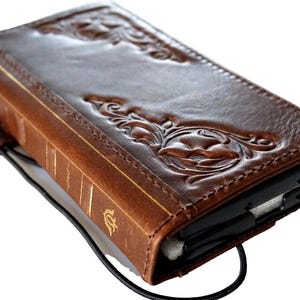 May include: Brown leather phone case with embossed floral design. The case has a book-like appearance with gold-colored accents on the spine. A black cord and a small leather tab are attached to the case.
