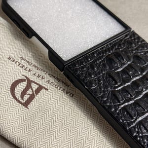 May include: A black phone case with a textured crocodile skin pattern on the back. The case features a black frame and a white foam insert. The case is placed on a beige fabric pouch with a herringbone pattern and the text DAVIDOV ART ATELIER.