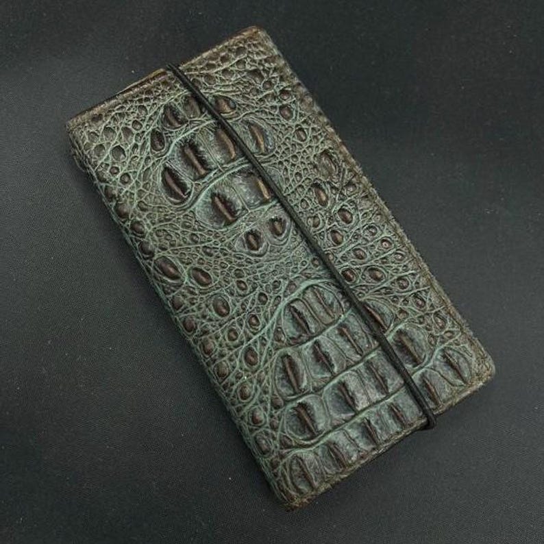 May include: A green and brown alligator skin wallet with a black elastic band closure.