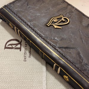 May include: A dark brown leather journal with a textured surface and a metallic Eye of Horus emblem on the cover. The spine features gold-colored line details and small decorative symbols. The text DAVIDOV ART Handmade is visible nearby.