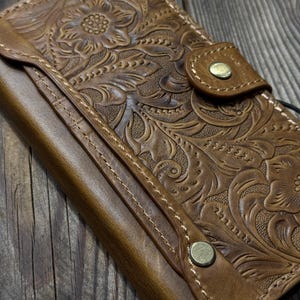 Puede incluir: Cartera de cuero marrón con diseño floral en relieve. La cartera presenta un cierre de botón a presión y costuras visibles. El cuero tiene un aspecto envejecido, con una estética vintage.