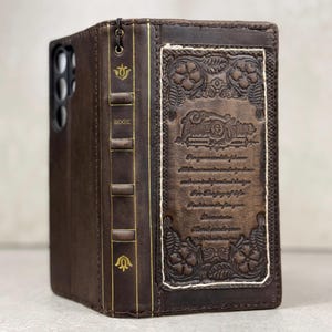 May include: A dark brown leather phone case designed to resemble an antique book. The spine features gold-stamped floral motifs and the word BOOK. The front cover displays an embossed floral border and intricate text including the year 1948.