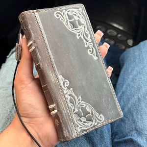 May include: A rectangular dark brown leather wallet case featuring white floral scrollwork patterns in the top and bottom corners. The spine has visible white stitching details and a thin black wrist strap attached to the side edge.
