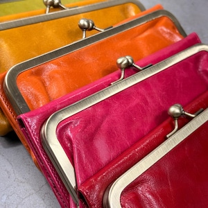May include: A collection of colorful leather clutch purses in shades of yellow, orange, pink, and red. Each purse features a metal clasp closure and a rectangular shape. The purses are arranged in a row, showcasing their vibrant colors and textures.