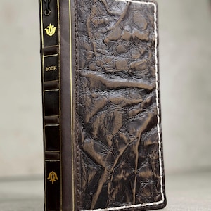 May include: A dark brown leather book cover with a textured surface and white stitching. The spine is dark brown with gold accents and the word "BOOK". The cover has a gold floral design.