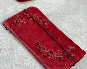 May include: A red leather wallet with an embossed floral design. The wallet is rectangular with a black elastic band closure. The leather has a textured appearance. A red leather strip and a crafting tool are in the background.