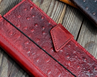 May include: A close-up of a red leather wallet with a textured surface and a black elastic band. The wallet has a flap closure and is stitched around the edges. Other leather wallets in brown and black are visible in the background.