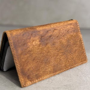 May include: A brown leather wallet, open to reveal a grey interior. The wallet has a distressed, textured appearance with visible stitching along the edges. The leather is a warm, earthy tone.