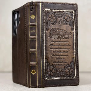 May include: A brown leather phone case designed to resemble a vintage book. It has gold detailing, embossed floral patterns, and text. The spine is labelled "BOOK" in gold. The case is designed to fit a smartphone.