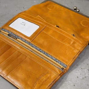 May include: Mustard-yellow leather wallet, open to reveal multiple compartments and a clasp closure. The interior includes card slots, a zipped pocket, and a patterned lining. A white tag reads "40017 Yellow".