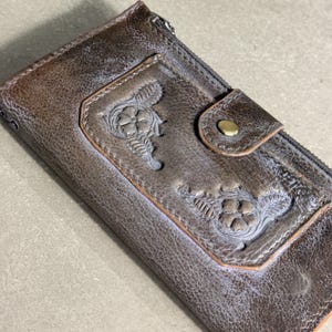 May include: A rectangular brown leather wallet featuring a distressed finish and a textured surface. The front panel displays two embossed floral motifs and a snap-button closure with a brass-colored metal stud. A zipper runs along the top edge.