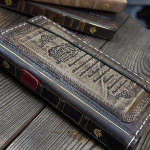 May include: A dark brown leather journal cover with a rectangular embossed center panel featuring ornate scrollwork and text. The spine has gold-colored decorative accents and a small red leather detail. A black elastic band secures the cover.