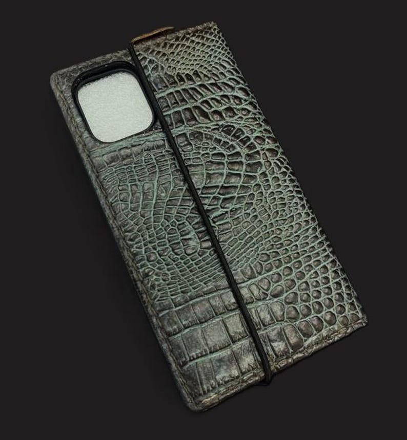 May include: A green crocodile-embossed leather phone case with a black elastic band closure.