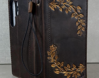 May include: Dark brown leather phone case with a gold-colored leaf design. The case has a strap and a cutout for the phone's camera. The case is designed to fold over and protect the phone's screen.