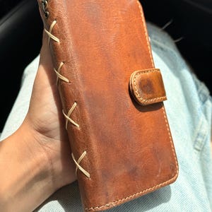 May include: A brown leather phone case with a rectangular shape and a magnetic closure flap. The side edge features decorative light-colored cross-stitching detail along the spine, providing a textured appearance to the smooth leather surface.