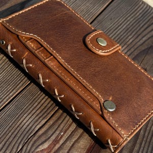 May include: A brown leather wallet with a button closure and decorative stitching. The wallet has a textured surface and a stitched design along the edges. The wallet is made of brown leather.