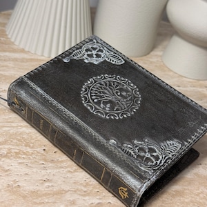 May include: A dark gray leather-bound book featuring an embossed tree of life design on the front cover, highlighted with silver-toned accents. The spine includes gold-colored horizontal lines and a small gold symbol at the bottom edge.