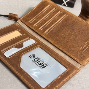 May include: A brown leather wallet featuring multiple card slots and a clear ID window with a white insert labeled RFID PROTECTION. The wallet has visible stitching along the edges and a fold-over design with a small tab closure on the side.