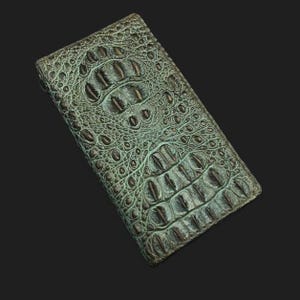 May include: A teal alligator skin wallet with a dark brown undertone. The wallet is closed and lying flat on a black surface.