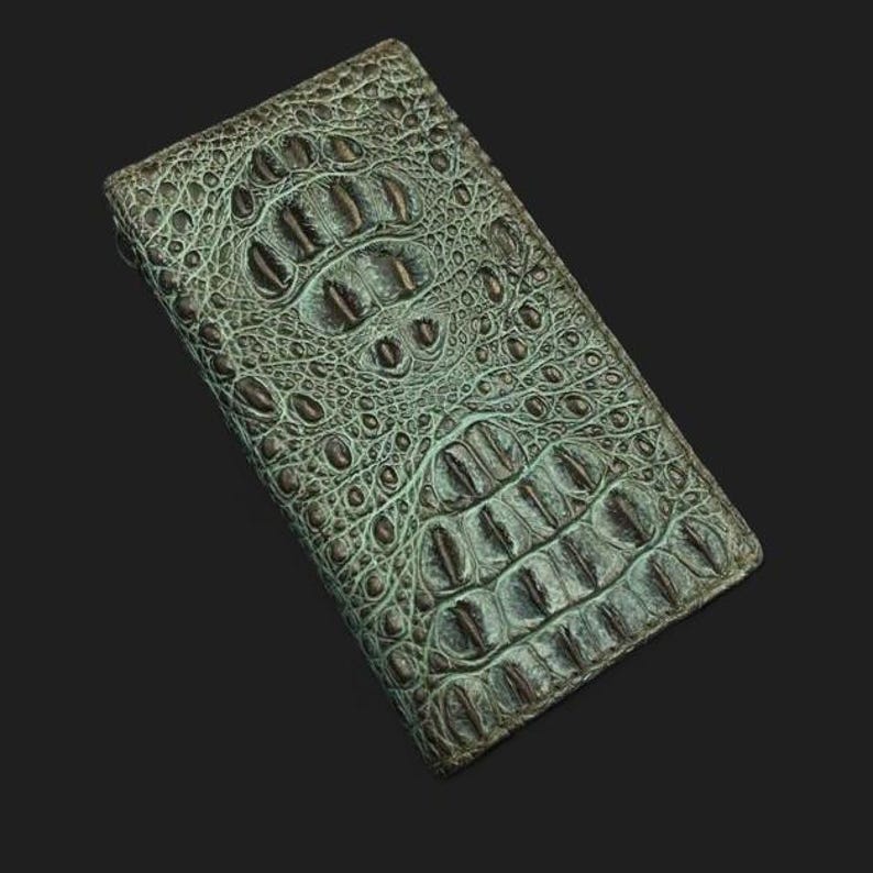 May include: A teal alligator skin wallet with a dark brown undertone. The wallet is closed and lying flat on a black surface.