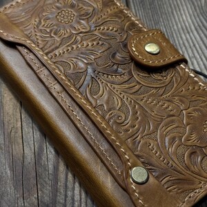 May include: A brown leather wallet with a floral embossed design. The wallet has a snap closure and visible stitching. The leather has a distressed look, and the wallet appears to be new.