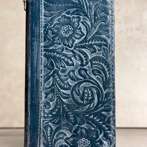 May include: A rectangular dark blue leather wallet featuring an intricate embossed floral pattern across the front surface. The design includes detailed carved flowers and swirling leaves with a distressed finish that highlights the texture.