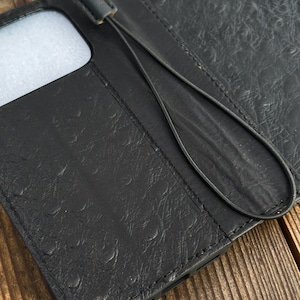 Puede incluir: Funda de teléfono de cuero negro con un patrón texturizado y una correa para la muñeca. La funda está abierta, revelando un interior blanco. La funda está sobre una superficie de madera.