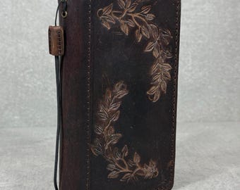 May include: Dark brown leather wallet with embossed leaf design. The wallet has a strap and a small leather loop attached. The leather has a distressed look. The wallet is rectangular and appears to be closed.