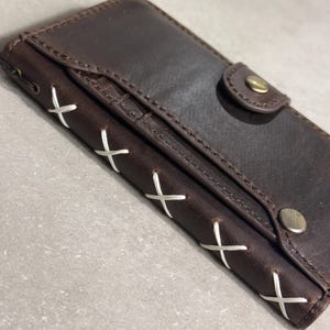May include: A dark brown leather wallet featuring a flap closure secured with a brass-toned snap button. The side edge is detailed with white cross-stitching, and an additional brass-toned snap button is visible on the front pocket flap.