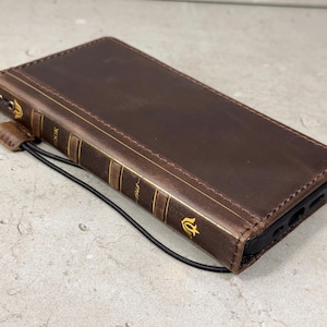 May include: A brown leather phone case designed to look like a vintage book. The case has gold-colored accents on the spine and edges, with a black elastic cord attached. The case is closed, showing the front cover.